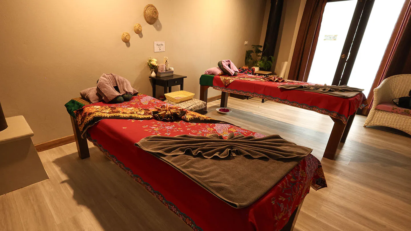 Two massage beds with decorative linens and towels, warm lighting, cultural accents, a wicker chair with a potted plant, and wood-toned flooring and walls for a relaxing ambiance