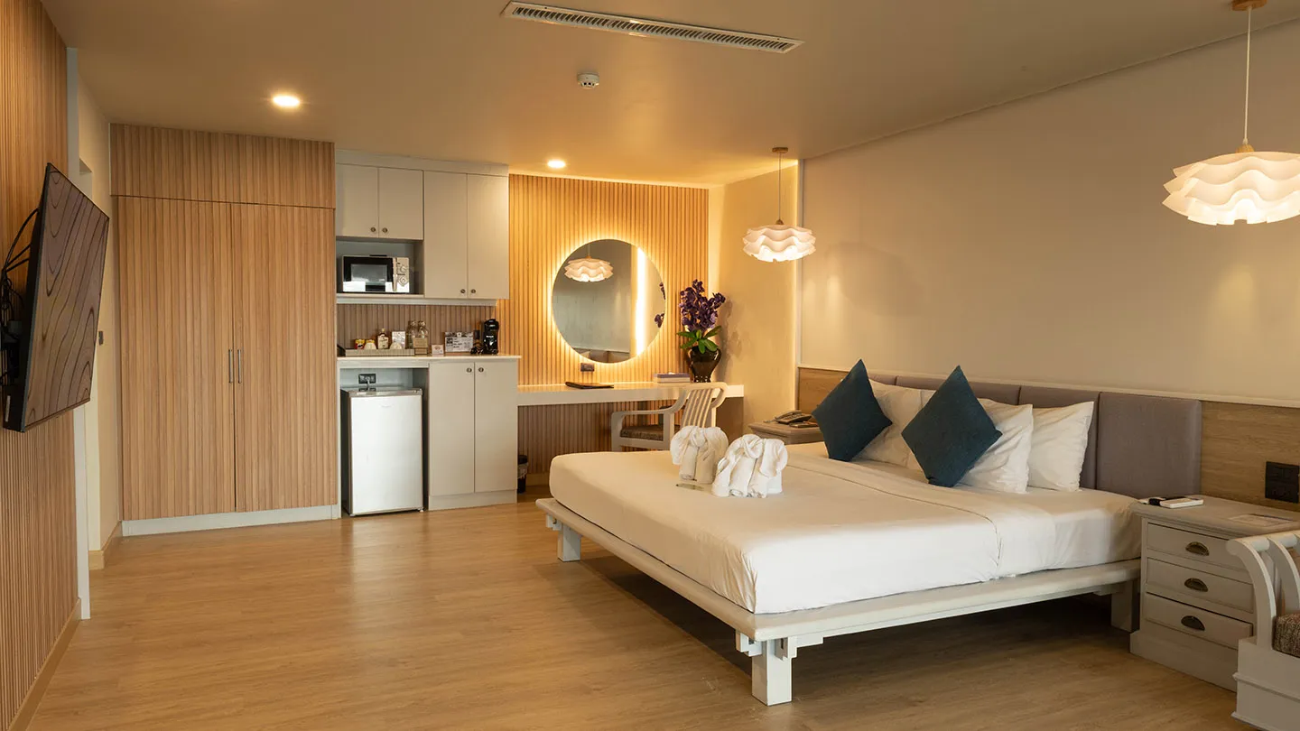 A spacious, modern room with a large bed by a window, light-colored walls, wooden accents, contemporary furniture, a dressing table, built-in storage, and warm lighting